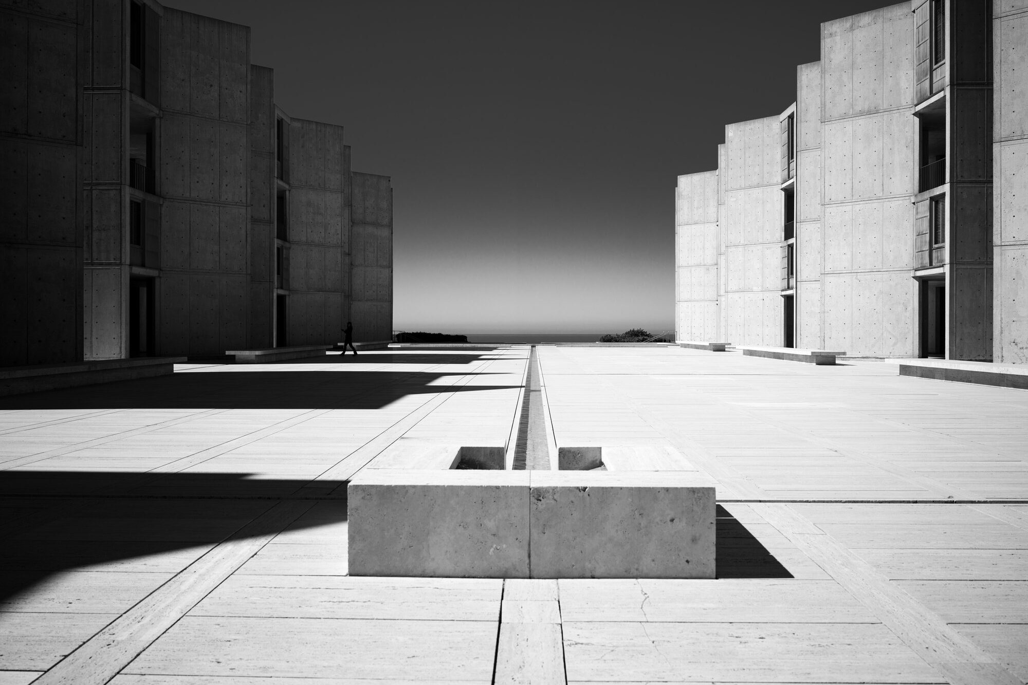 The Salk Institute plaza from its east end: the travertine court, the thin water channel running west to the Pacific, the study towers stepping down both sides, one figure walking at the left.