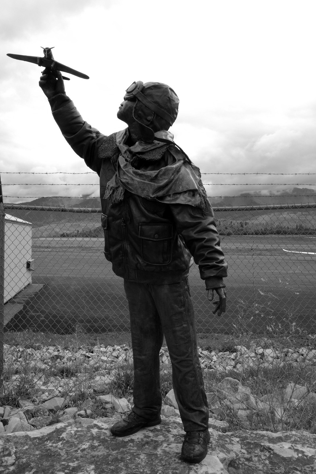 A bronze statue of a boy in flying goggles holds a toy plane above an airfield fence.