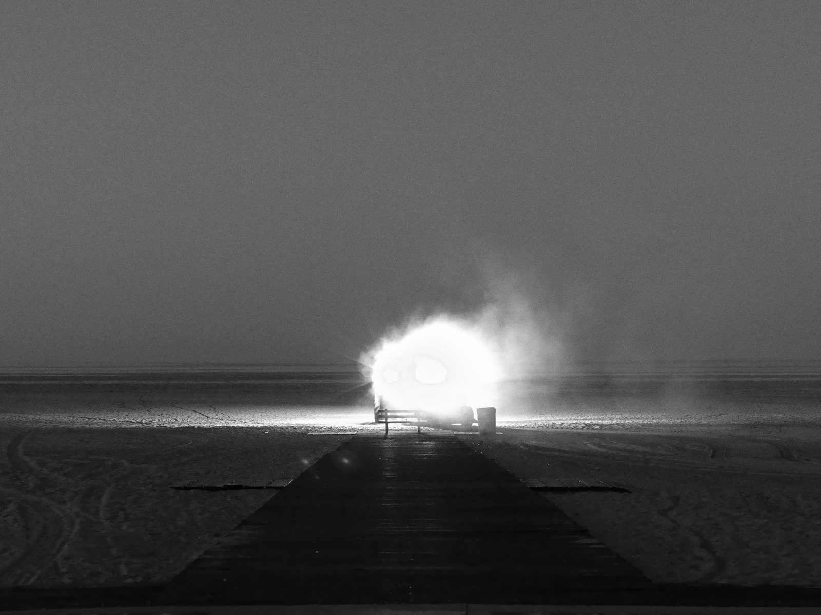 A beach-grooming machine glows under floodlights on dark sand at dawn, a boardwalk leading toward it.