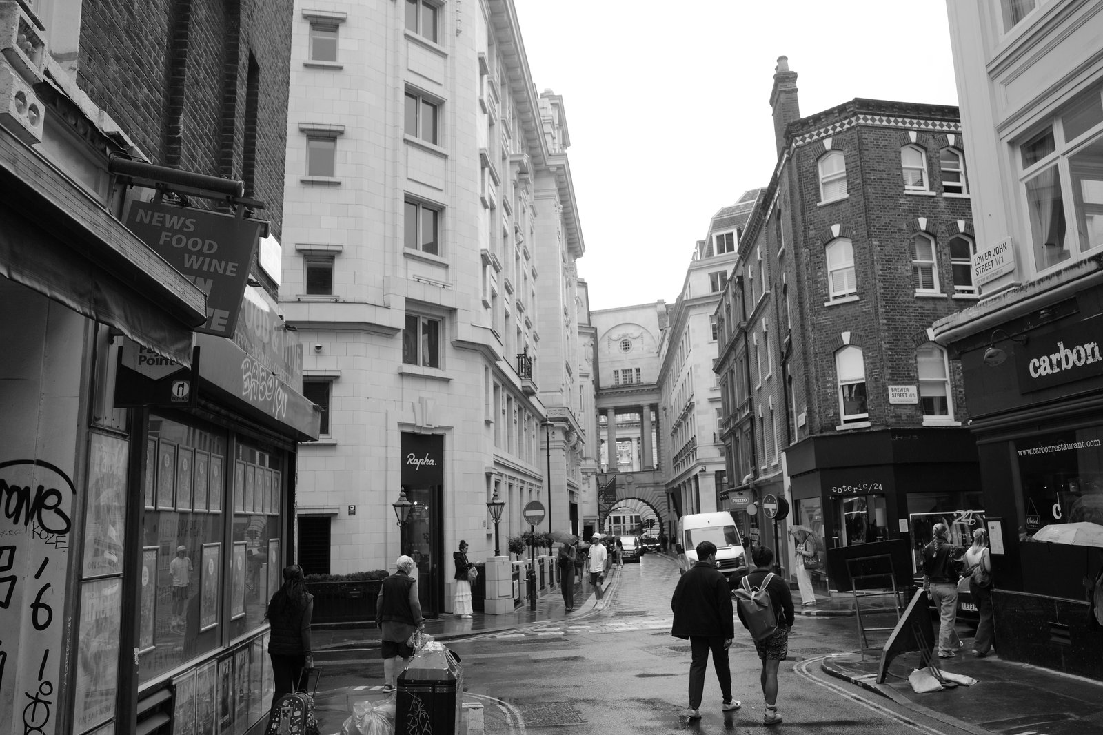 Pedestrians walk between layered facades along a narrow wet street.