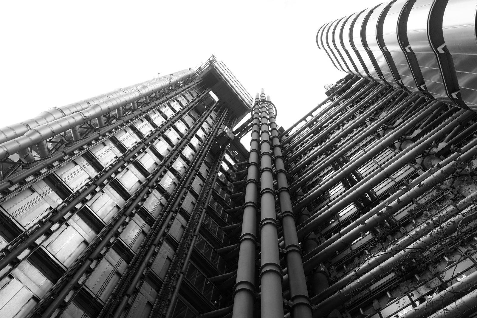 Rows of exposed service pipes converge overhead between building facades.