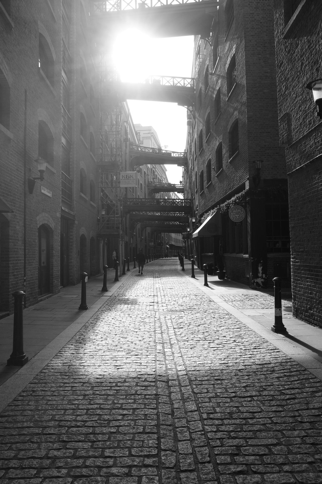 Sunlight falls across a cobbled passage between dark walls and overhead bridges.