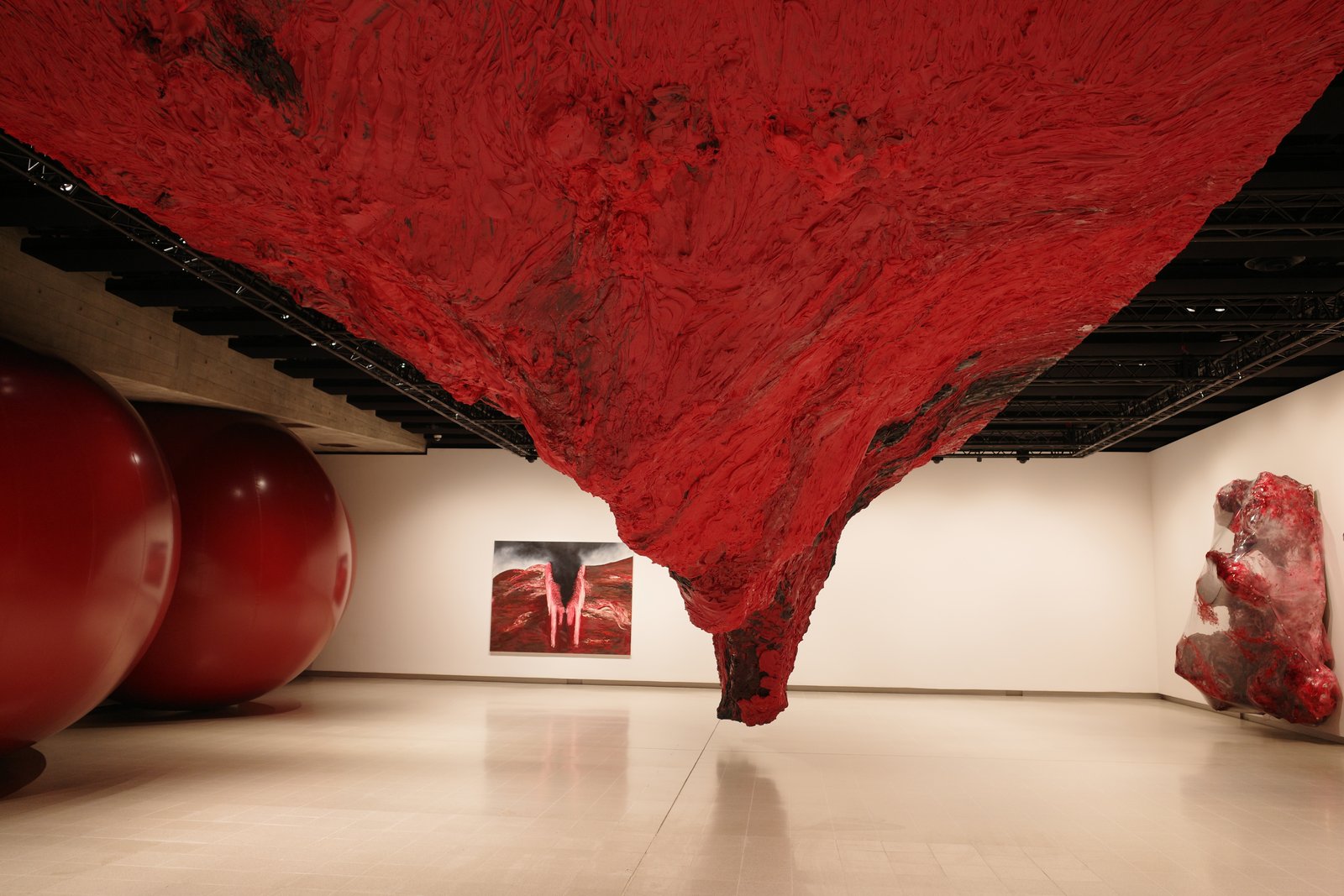 A red sculptural mass hangs above a gallery floor near other rounded forms.