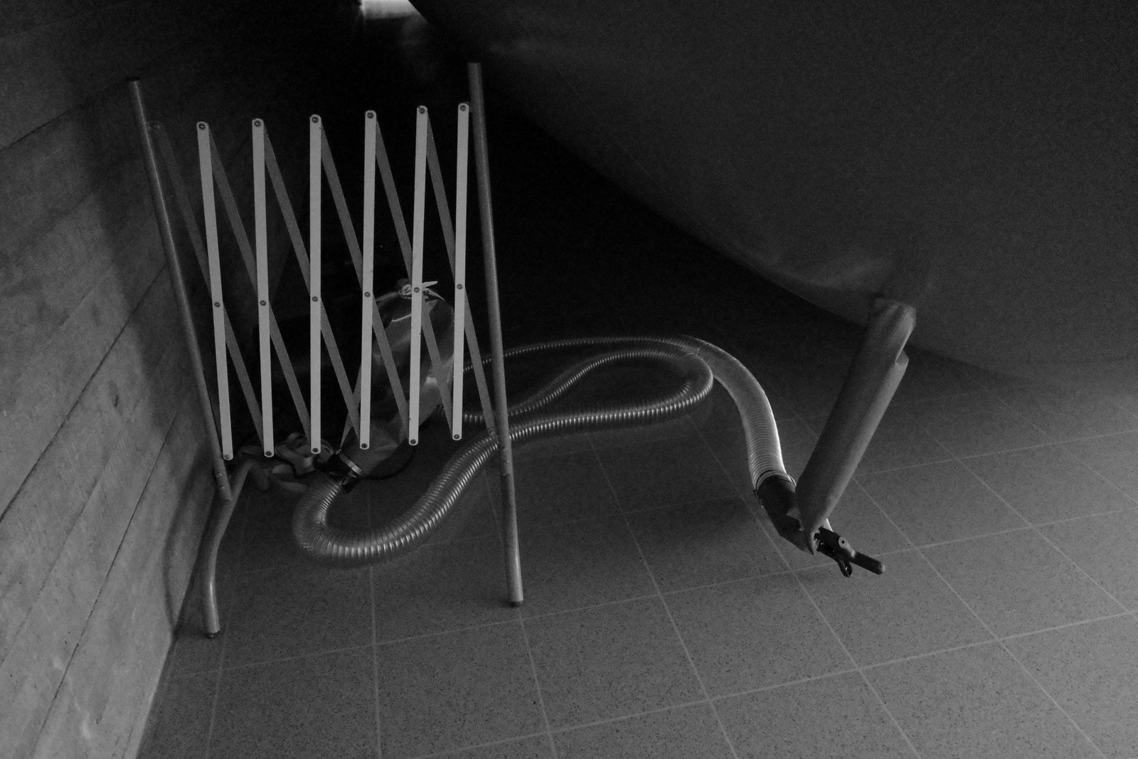 A hose and folding barrier rest on a tiled floor beside a dark curved volume.