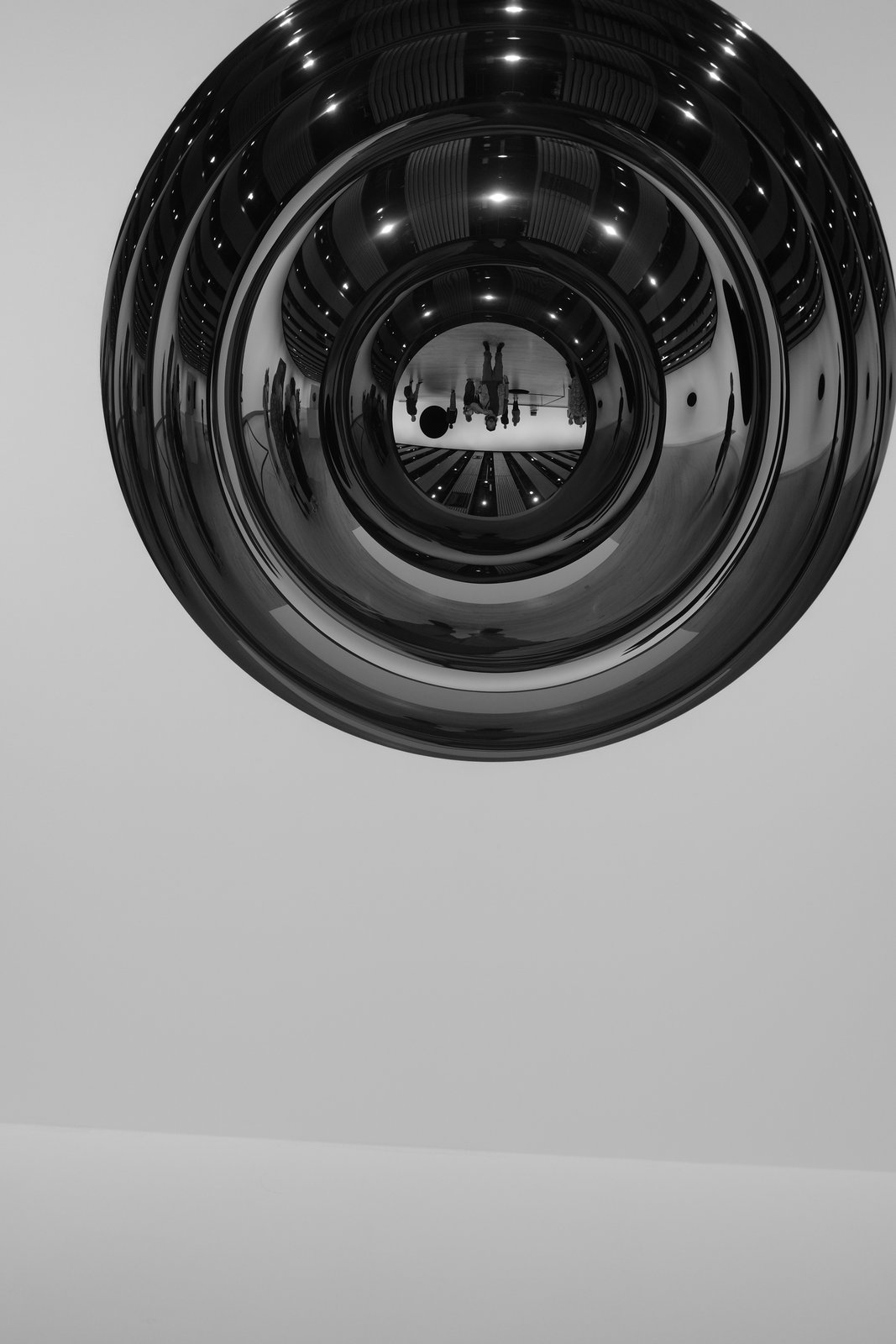 A reflective circular object contains concentric reflections of a room and its visitors.