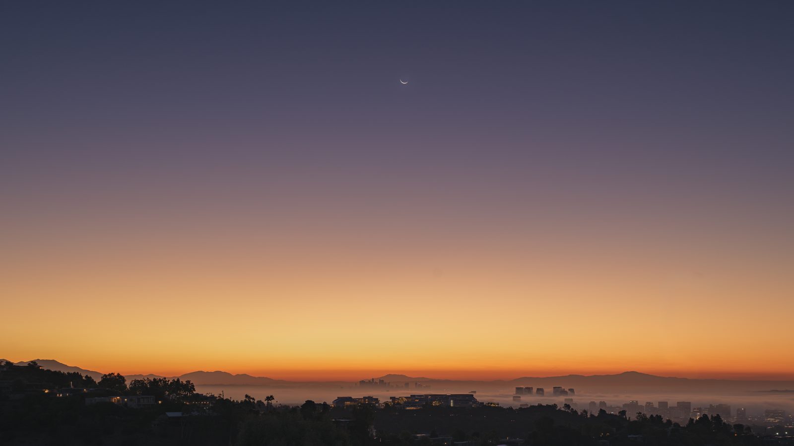 A thin crescent moon hangs over a city skyline at dawn in an orange and violet sky.