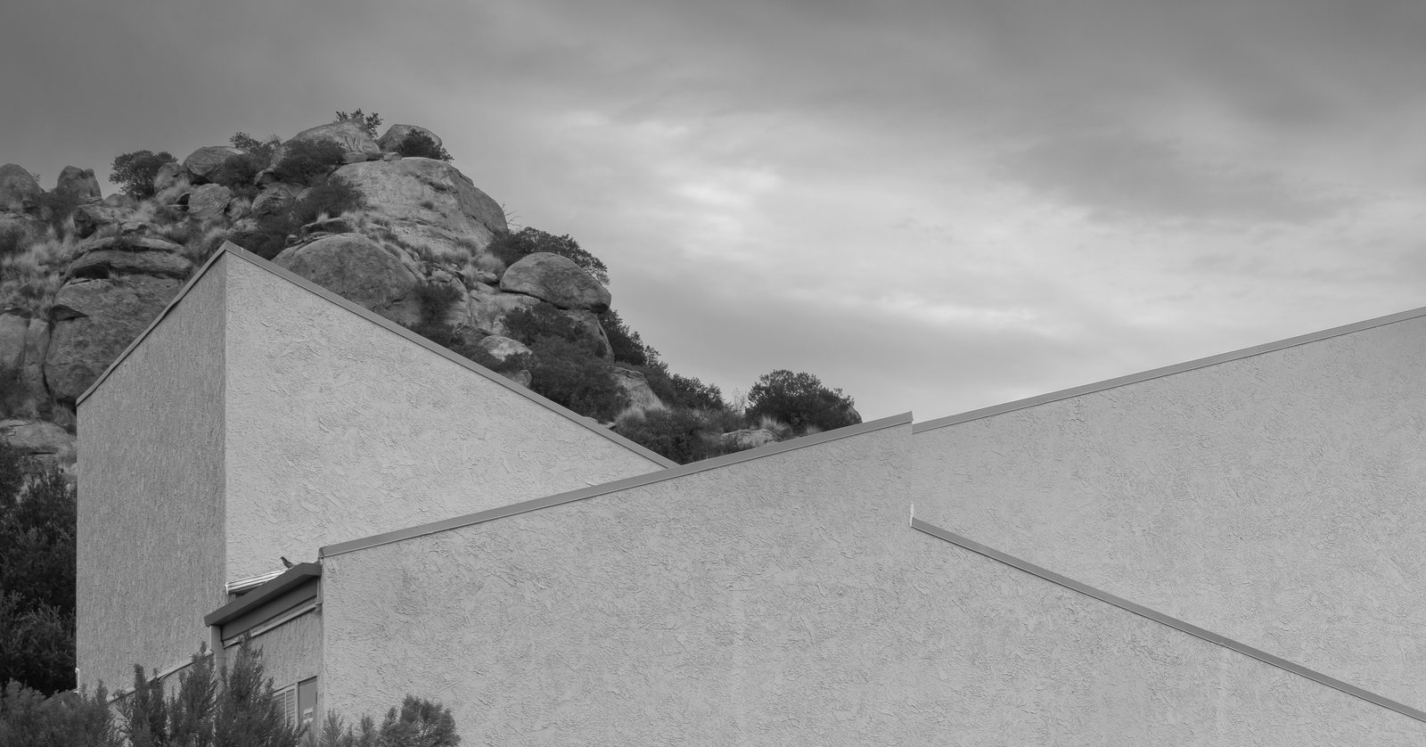 Two pale stucco roofs angle against each other below a rocky hillside under an overcast sky.