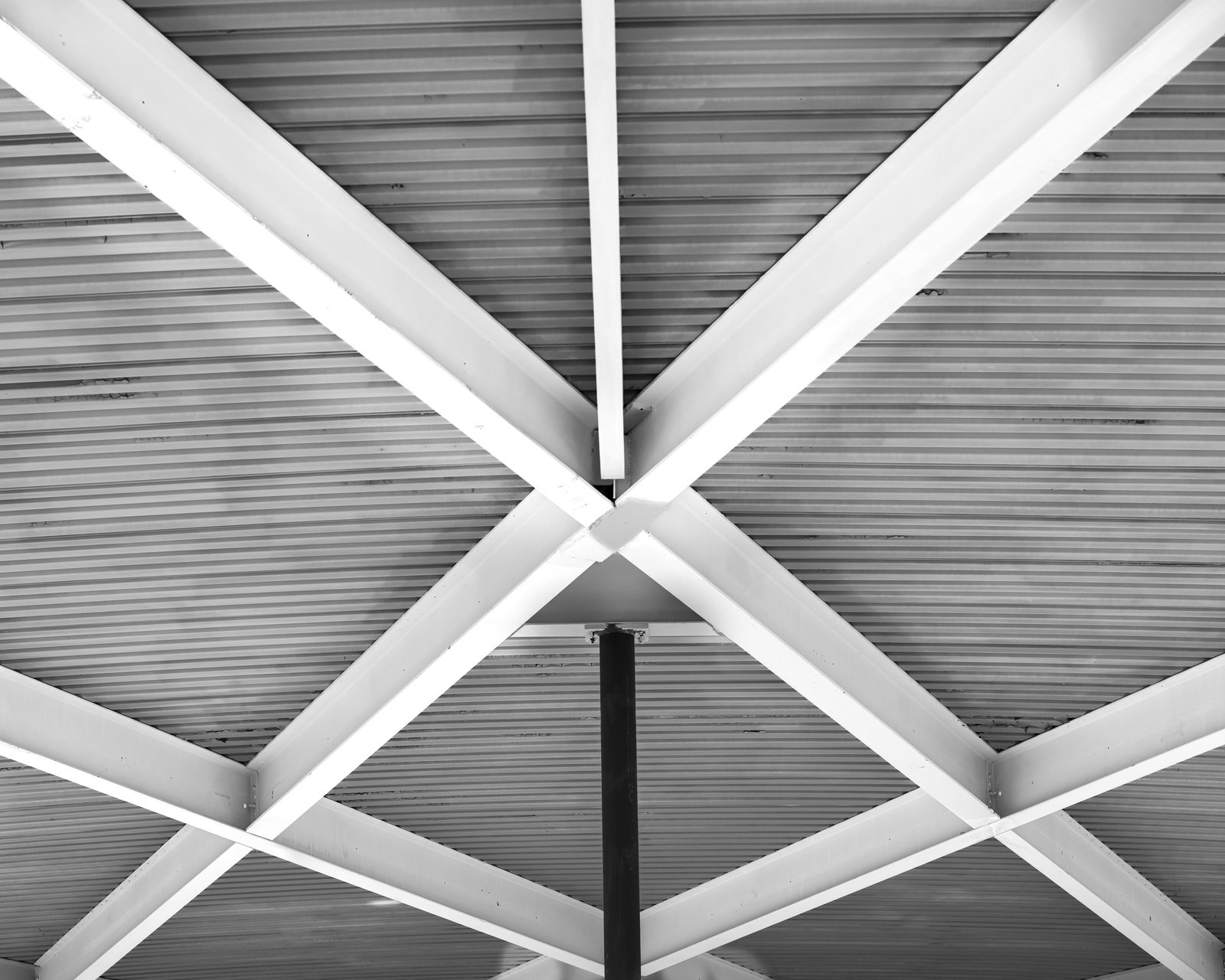White steel beams cross in an X beneath a corrugated ceiling above a dark column.