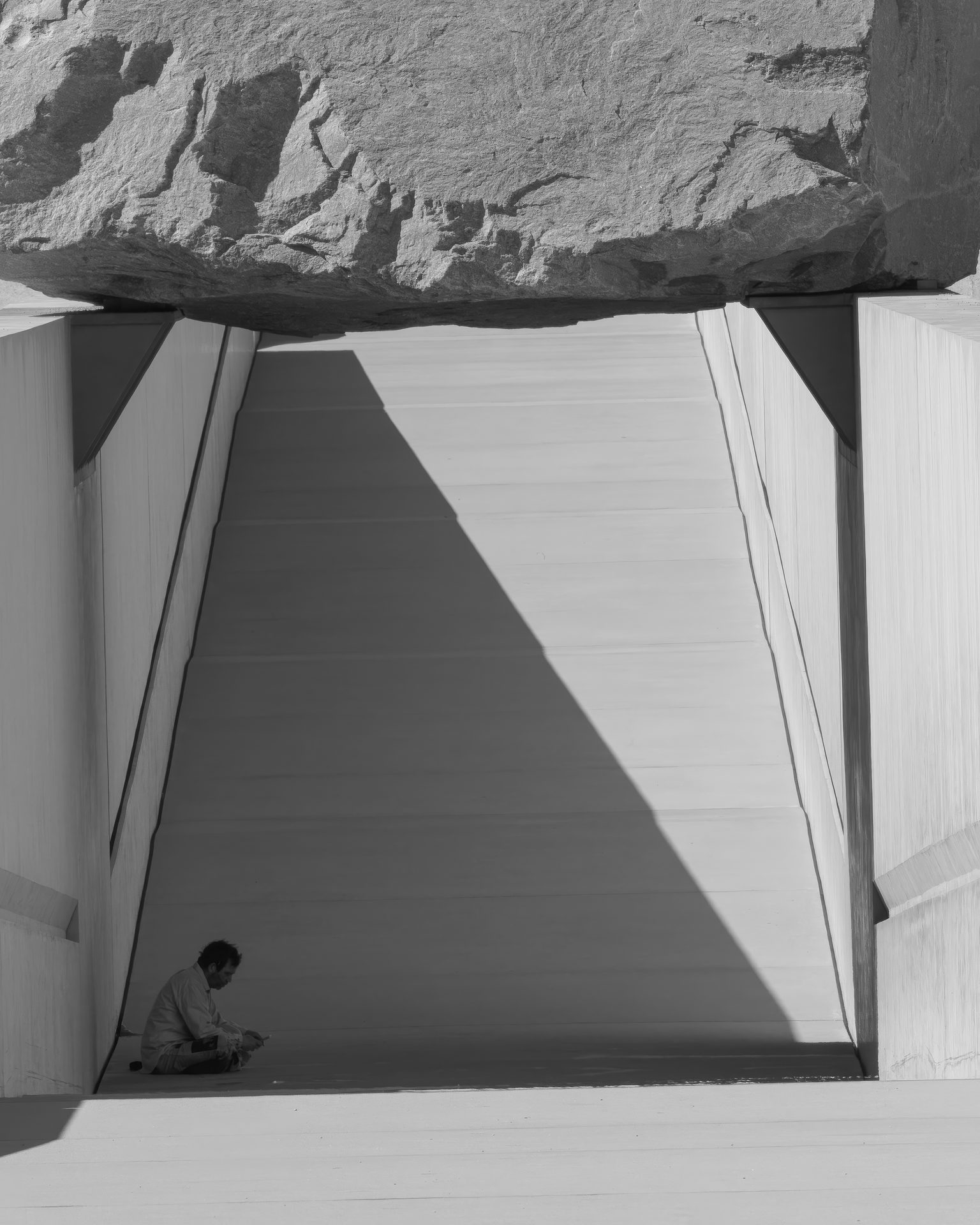 A person sits at the foot of a sunken concrete walkway beneath a massive boulder spanning it.