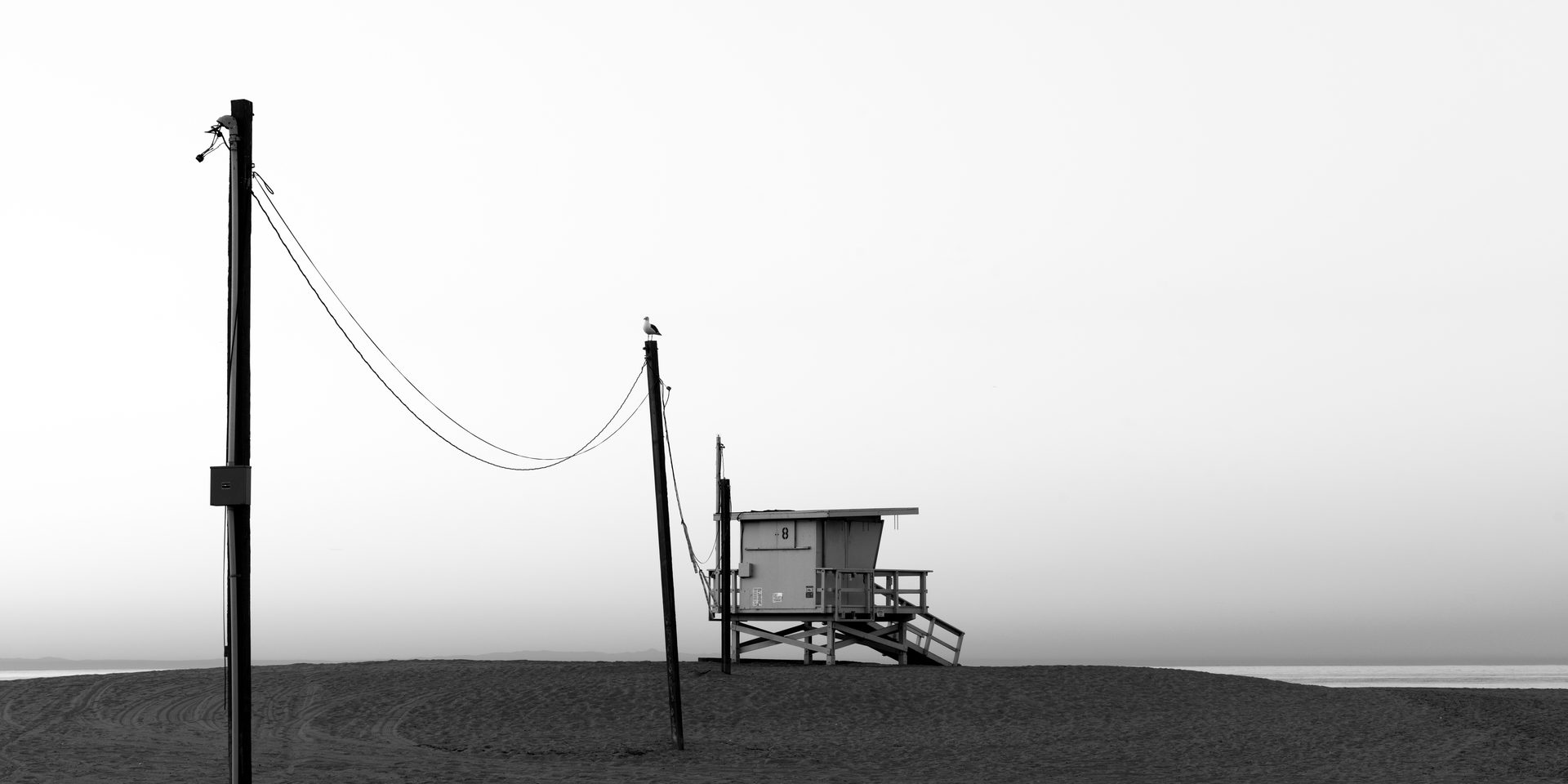 A lifeguard tower stands on a bare beach between two wired poles under a pale sky.