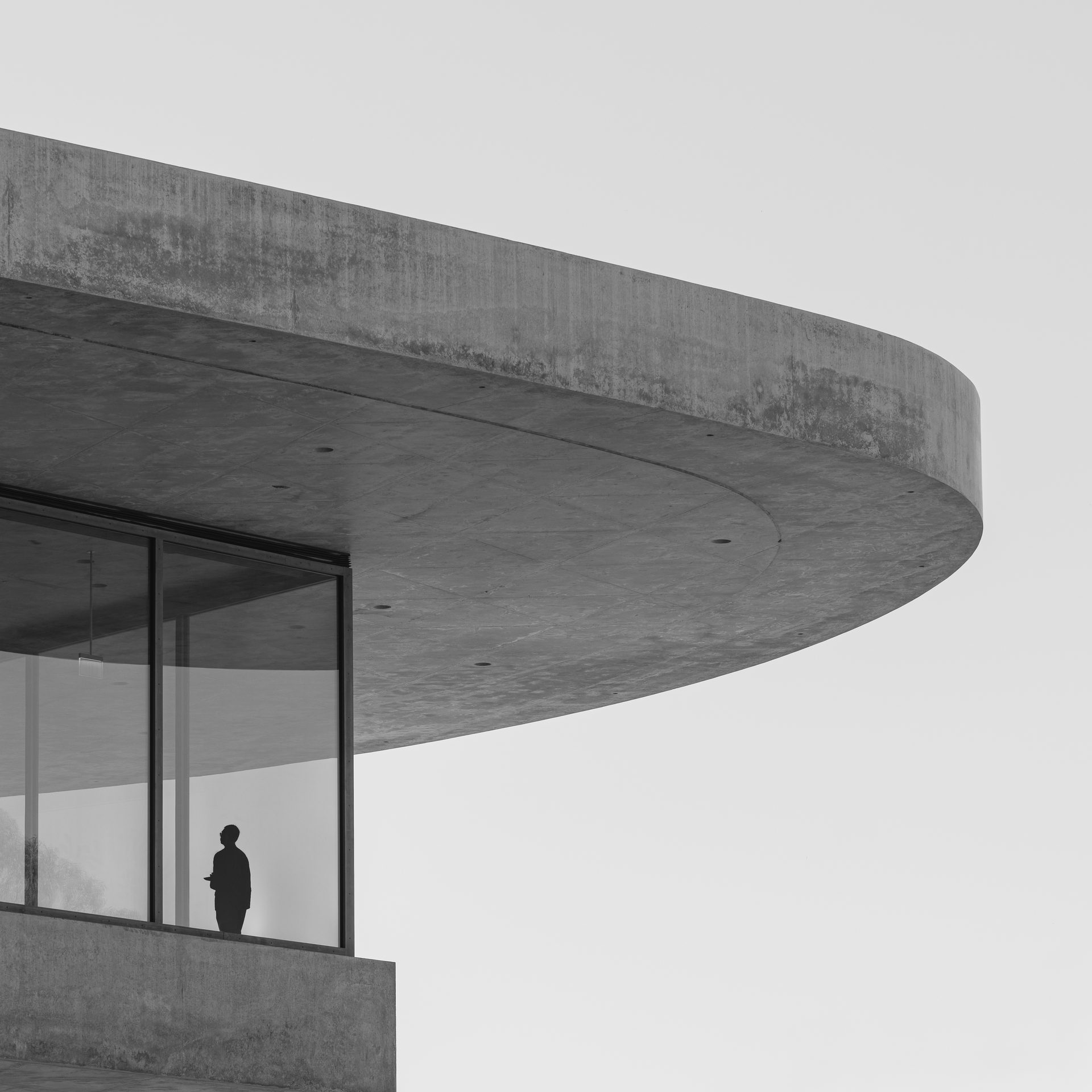 A lone figure stands behind glass beneath a wide concrete disc that overhangs the building.