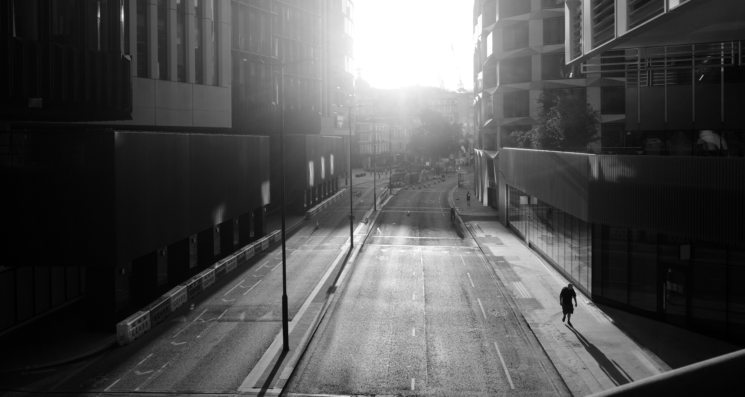 Low sun shines down a street between tall buildings, beside a lone walker.