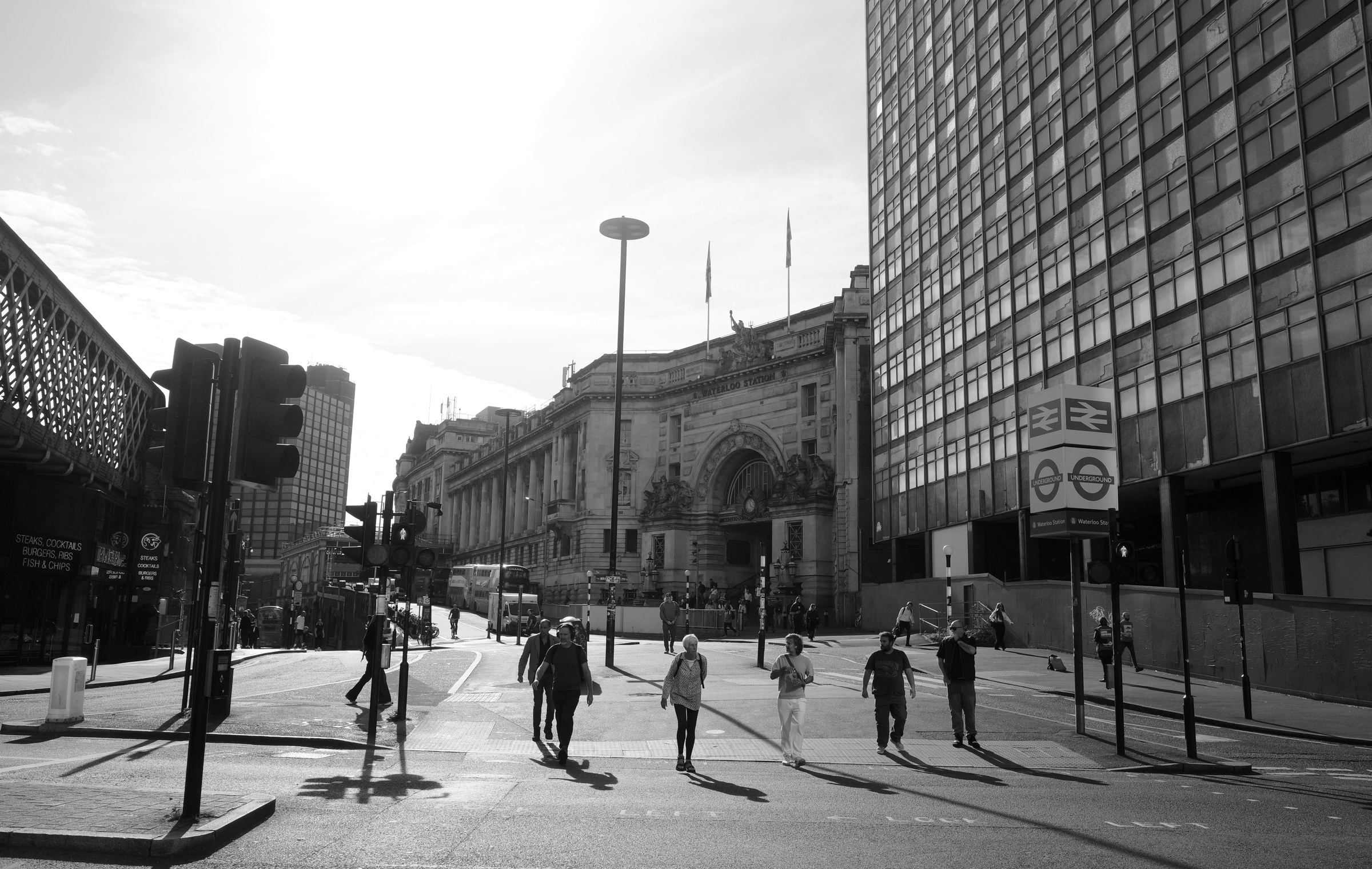 Pedestrians cross a sunlit street in front of Waterloo Station.